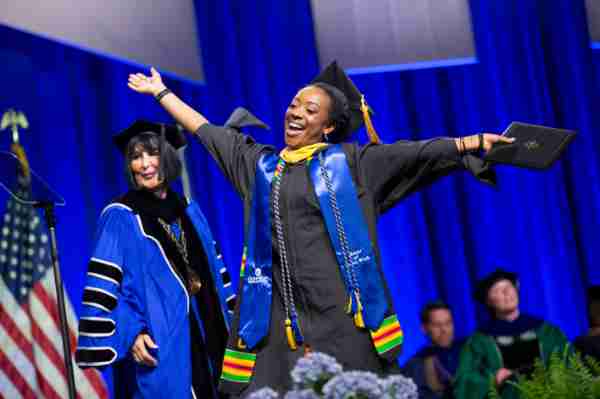 A grad throws her arms out in triumph in front of President Mantella as she walks across the stage.