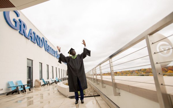 A person wearing a cap and gown has their hands in the air. The words Grand Valley State are on a building in the background.