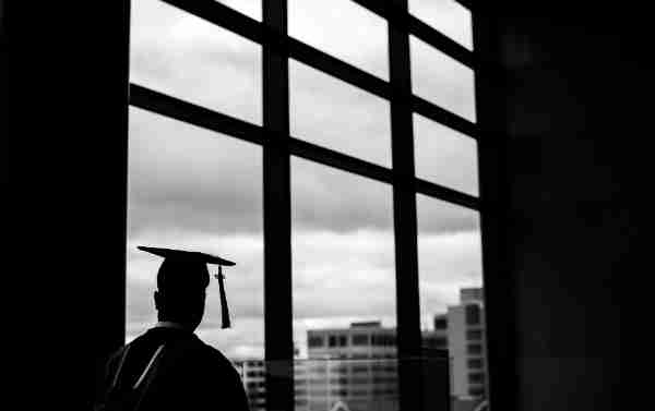 A person wearing a cap and a tassel looks out a window at buildings.