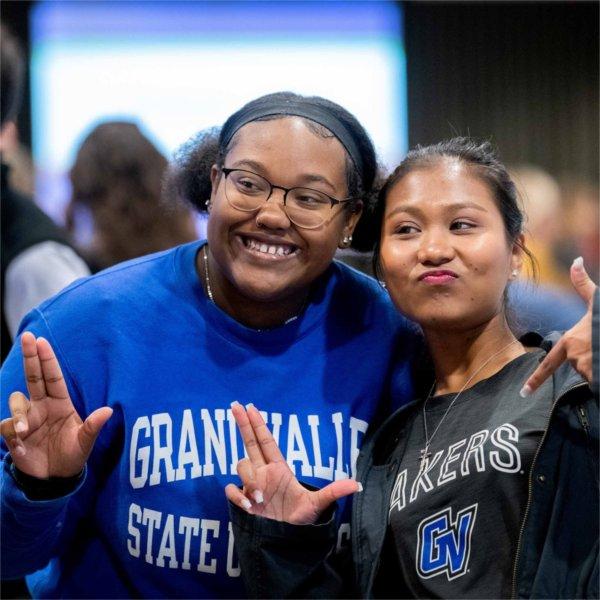 Two students in Grand Valley shirts smile together and hold their fingers and thumb in an Anchor Up pose.