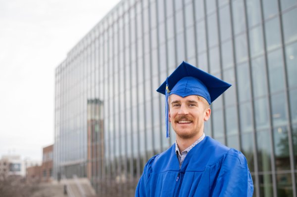 A person wearing a cap and gown smiles. The carillon is reflected off a building in the background.