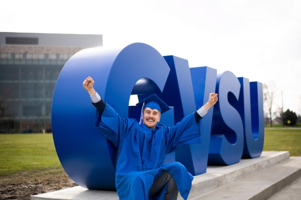 A person wearing a cap and gown smiles while pumping fists into the air with the letters GVSU in the background.