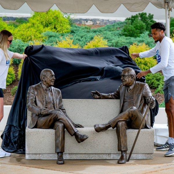 Two people pull a covering off a sculpture showing two people sitting on a bench.