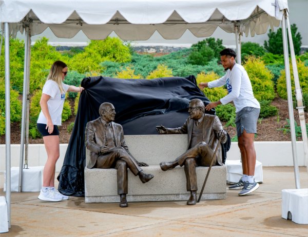 Two people remove a covering to reveal a sculpture of two people sitting on a bench.