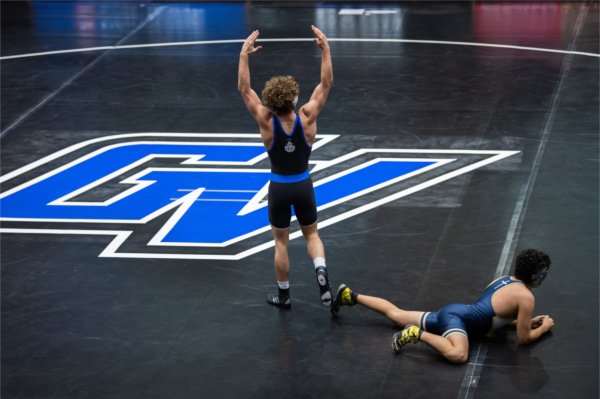 A person stands on the mat with his hands in the air in victory after a wrestling match while his opponent is still on the ground. 