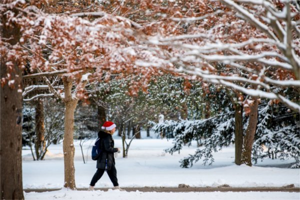A person walks on a sidewalk in snowy surroundings wearing a Santa hat.  