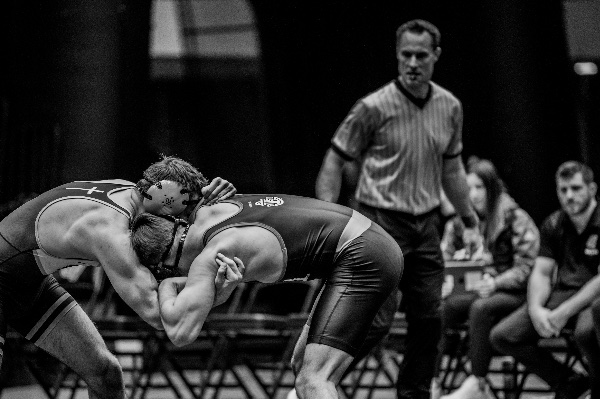 Two wrestlers are crouched with heads and arms entangled while a referee watches from behind them. 