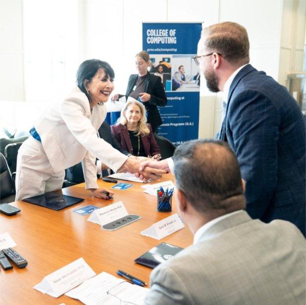 President Mantella leans over table to shake hands with Henry Ford College President David M. Knezek. A GVSU College of Computing vertical banner is in the background