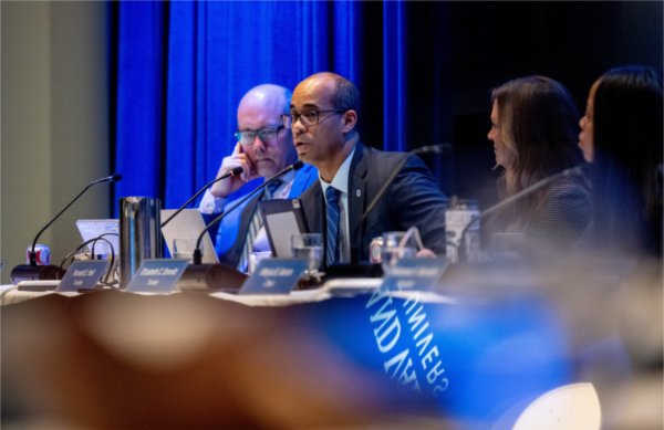 Grand Valley State University trustees listen during a Board of Trustees meeting, with a trustee speaking into a microphone at the conference table as other board members look on.