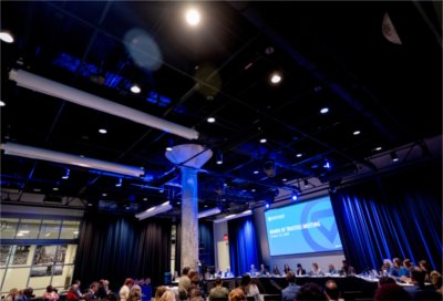 Grand Valley State University&#8217;s Board of Trustees meeting in a conference room, with trustees seated at a long table beneath blue stage lighting and a large screen displaying the meeting title.