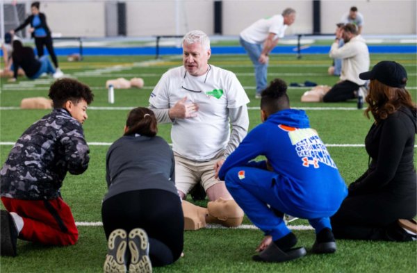 A person kneeling on the ground with a manikin before them talks to four others who are also kneeling.