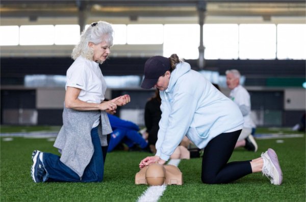 One person practices CPR on a manikin while another looks on. Both are kneeling on the floor.