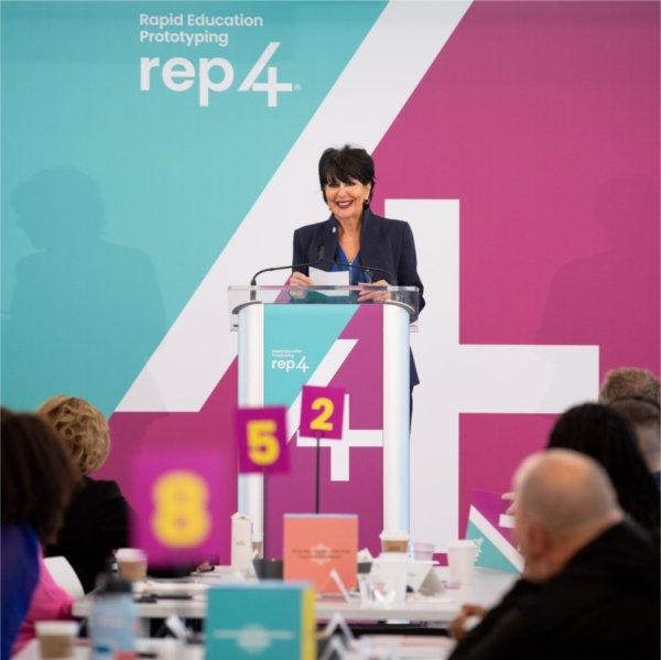 President Mantella stands in front of a turquoise and purple REP4 backdrop and addresses a room full of people.