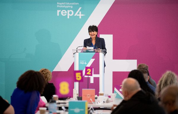 President Mantella stands in front of a turquoise and purple REP4 backdrop and addresses a room full of people. 