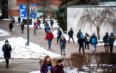 students walk on sidewalk, snow on grass areas; banner on lightpost reads Next is Now