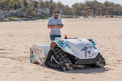 A man wearing sunglasses and a gray polo shirt stands on a sandy beach while operating a remote control for a small, tracked beach-cleaning robot.