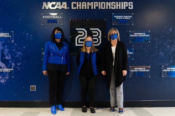 from left are President Mantella, Joan Boand and Keri Becker standing against a wall in the Fieldhouse that reads NCAA Championships