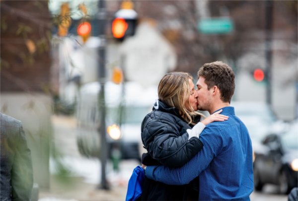 A couple kiss as they stand outside in a winter city scene. 