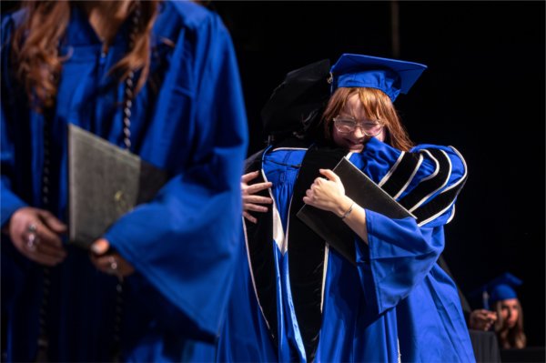 A graduate wearing a blue cap and gown hugs a university president after receiving their diploma. 
