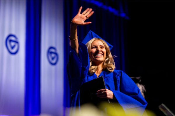  A smiling graduate wearing a blue cap and gown waves. 