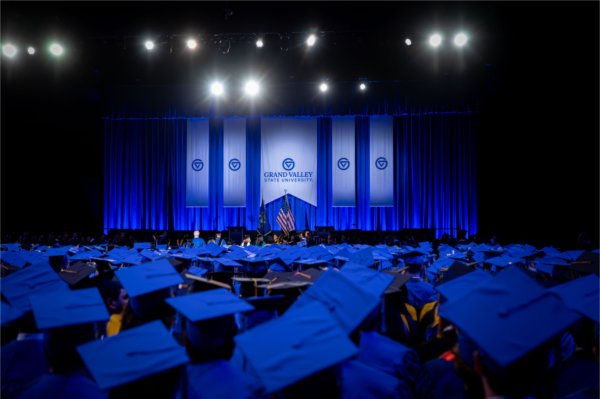  A large gathering of graduates wearing blue caps and gowns sit in an arena in front of banners and lights. 