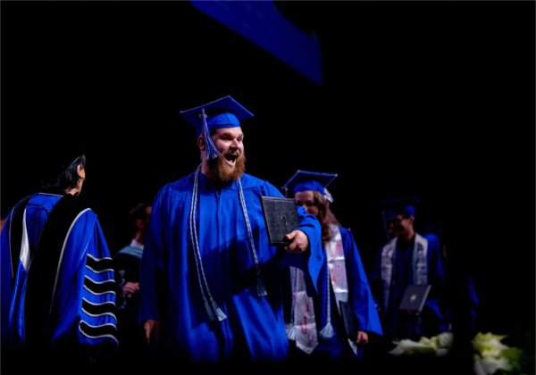  A graduate wearing a blue cap and gown makes a happy gesture as they cross the stage with their diploma. 
