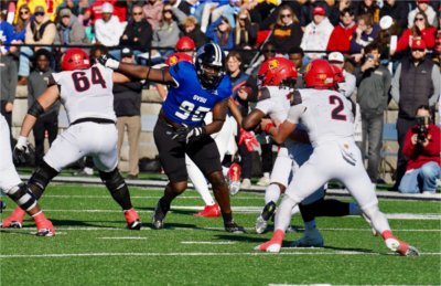 A GVSU defensive lineman in a blue jersey, number 95, pushes past an opposing blocker in white while the other team&#8217;s quarterback hands off the ball, with a crowd watching from the stands.