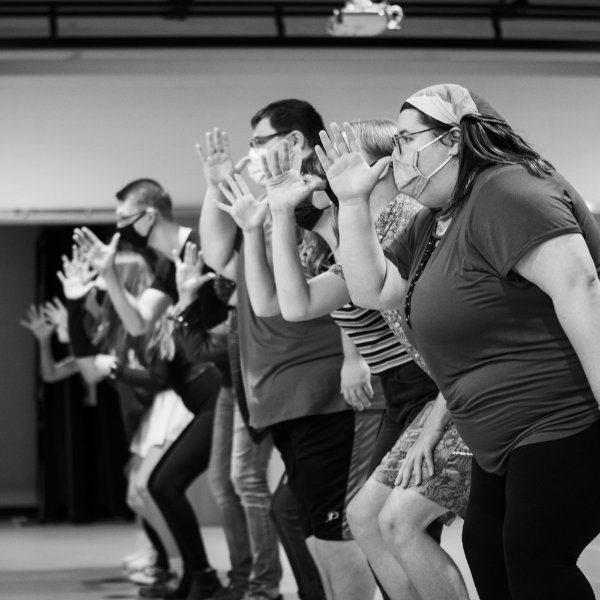 Performers at a rehearsal stand in a line with their hands in air as they sing.