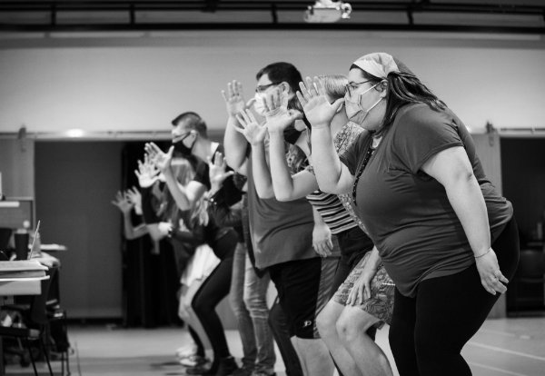 Performers at a rehearsal stand in a line with their arms in the air while singing.
