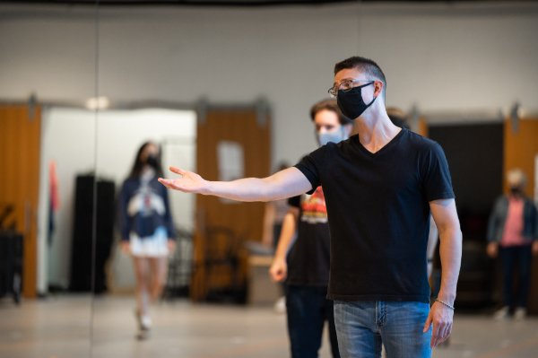A performer gestures with an outstretched right arm while singing. Other cast members are in the background.