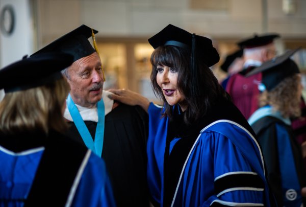 President Mantella puts her hand on Matthew McLogan's shoulder while talking to someone else. Both are in academic regalia.