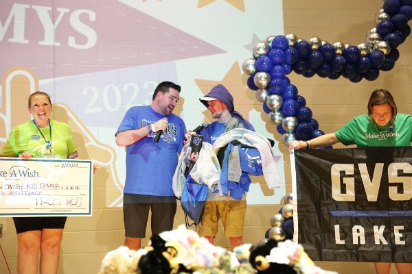 Connor Pung stands on stage holding a pile of GVSU clothing. Michael Hull stands next to him with a microphone, and two volunteers hold up a giant check and a GVSU Lakers flag.
