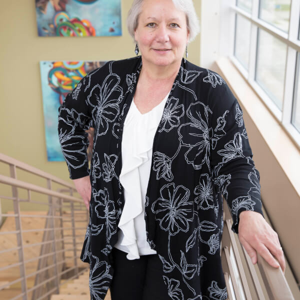 A portrait of Teri Behrens. She is standing at the top of a staircase in the Bicycle Factory Building, which houses the Johnson Center offices. There are several brightly colored abstract paintings on the wall behind her.