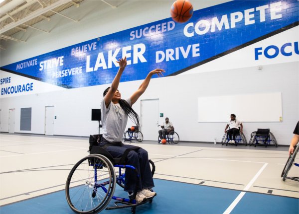  Student Kaliyah Brown sits in a wheelchair and shoots a basketball, with others in wheelchairs watching in the background.