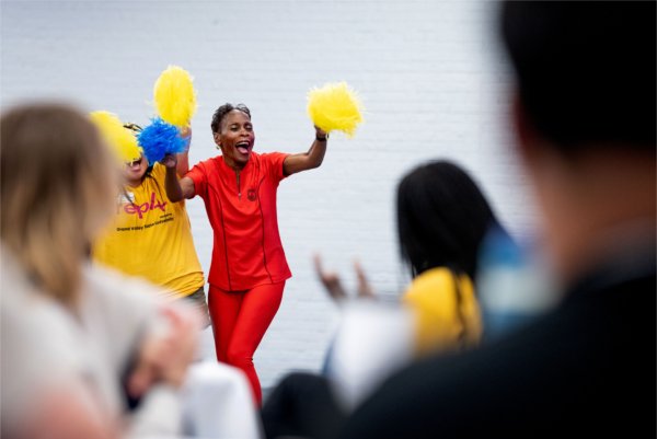 Two adults enthusiastically cheer with blue and yellow pom-poms in front of an audience, one wearing a red outfit and the other a yellow shirt. 