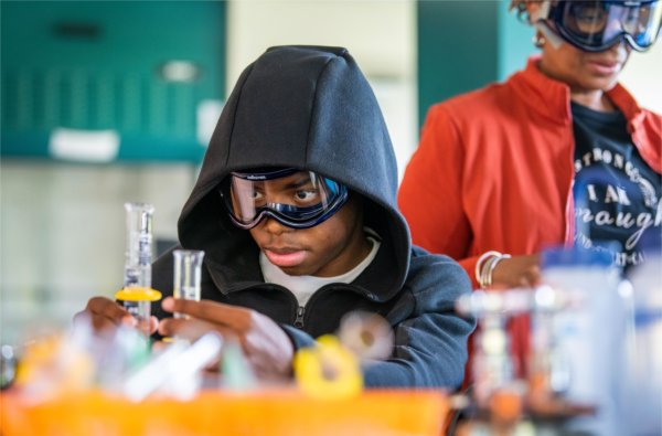 A student in a hoodie and safety goggles carefully examines a beaker in Padnos Hall of Science, with a supervising adult in the background.
