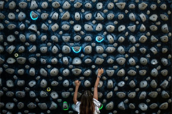 A young student reaches for handholds on a densely packed indoor climbing wall, with several holds lit by blue and green lights.