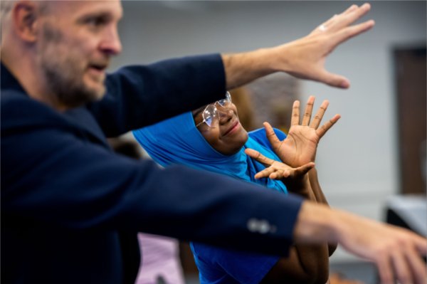 A smiling student in a blue hijab and glasses gestures expressively as an instructor in a navy blazer gestures beside her. 