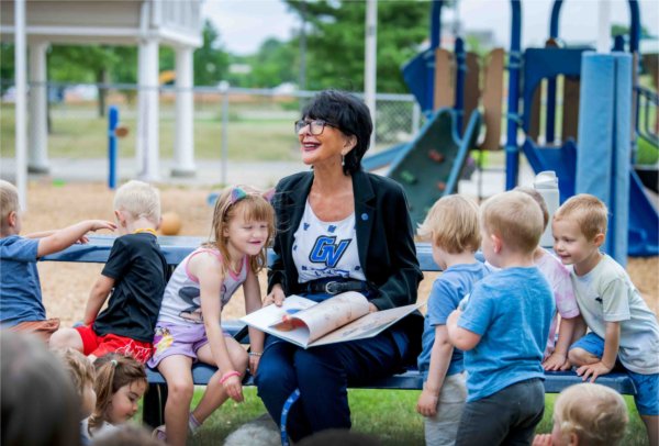 President Philomena V. Mantella sits on a bench smiling, as she holds a book open in her lap. Children sit beside her on the bench. 