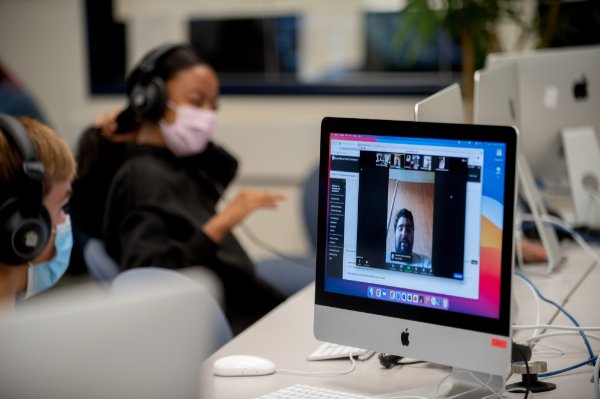 computer with student on screen in forefront, GVSU student in back with headphones on and a pink mask