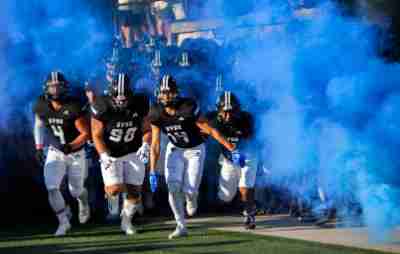 The Grand Valley football team runs on to the field through a cloud of blue smoke at the start of a game.