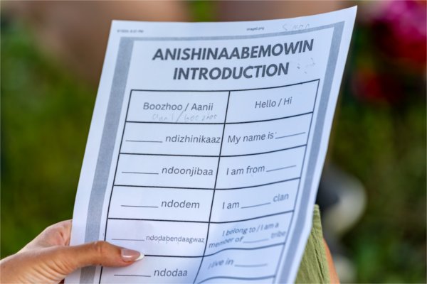 A hand holds up a paper that reads Anishinaabemowin Introduction, followed by a list of introduction sentences in both Anishinaabemowin and English, including &#8220;hello,&#8221; &#8220;my name is,&#8221; and &#8220;I live in&#8221;. 