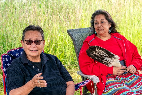 Two older women, one wearing a black polo, the other wearing a red sweatshirt and blue and red skirt, sit in lawn chairs. The woman in the skirt holds a large brown feather.