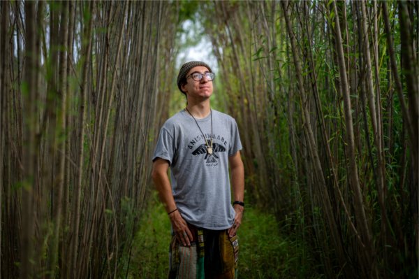 A young man wearing a beanie and grey shirt that reads &#8220;Anishinaabe&#8221; stands in a small clearing between two rows of thin trees. 