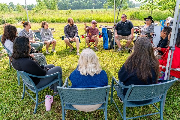 People sit in a circle of lawn chairs in a green grass field. One young man wearing a maroon beanie points as he talks.