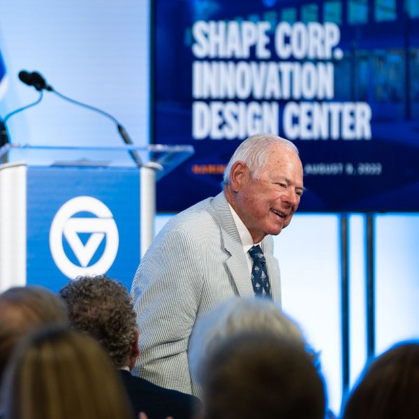 Midge Verplank, co-founder of Shape Corp. stands to be recognized at an event. In the background a screen shows Shape Corp. Innovation Design Center