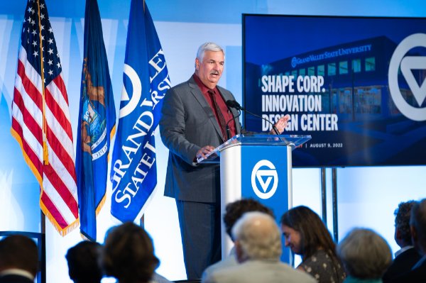 Paul Plotkowski stands on stage behind podium, screen reads Shape Corp Innovation Design Center