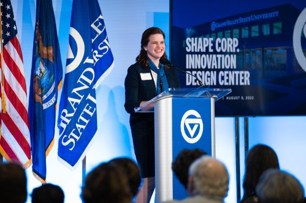 Vice President Laura Aikens stands behind a podium, screen reads Shape Corp Innovation Design Center