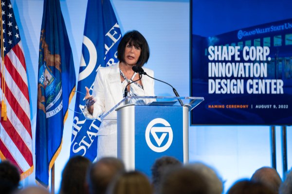 President Mantella speaks behind podium, flags behind her and a screen that reads shape Corp Innovation Design Center
