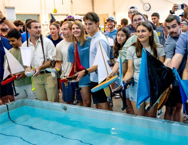 students holding sailboats stand around the edge of a shallow pool